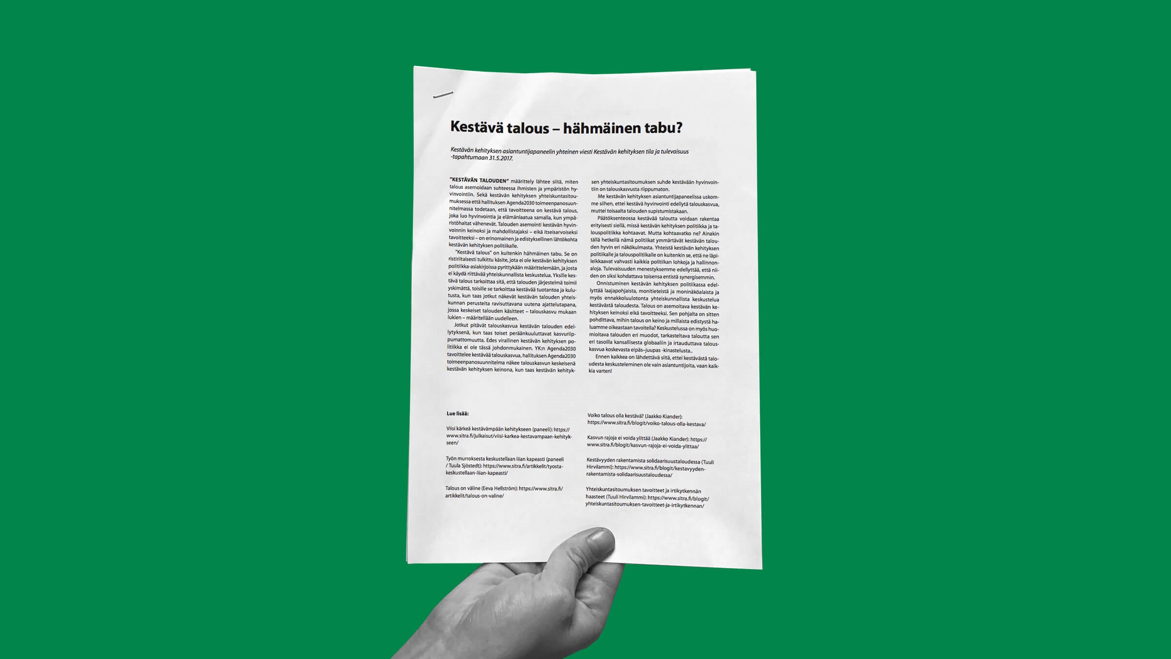 Käsi nostaa ylös painettua asiakirjaa, jossa on suomenkielistä tekstiä vihreää taustaa vasten. Asiakirjan yläosassa on lihavoitu otsikko Kestävä talous - hämäläinen tabu?.