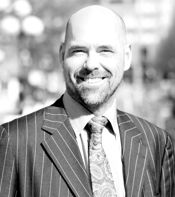 A smiling bald man with a beard is wearing a pinstripe suit, patterned tie, and collared shirt. The photo is in black and white, and the background is blurred, suggesting an outdoor urban setting.