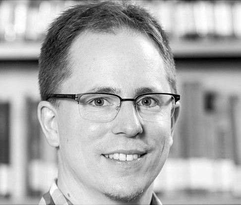 A man with short hair and glasses smiles at the camera. He is wearing a plaid shirt. The background is blurred but shows shelves filled with books. The image is in black and white.