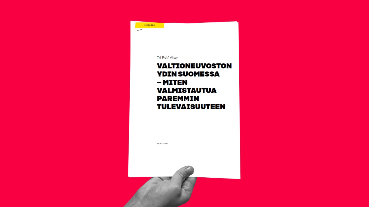Käsi pitelee valkoista vihkosta, jonka kannessa on suomenkielinen teksti kirkkaan vaaleanpunaista taustaa vasten. Otsikossa lukee: VALTIONEUVOSTON YDIN SUOMESSA - MITEN VALMISTAUTUA PAREMMIN TULEVAISUUTEEN.