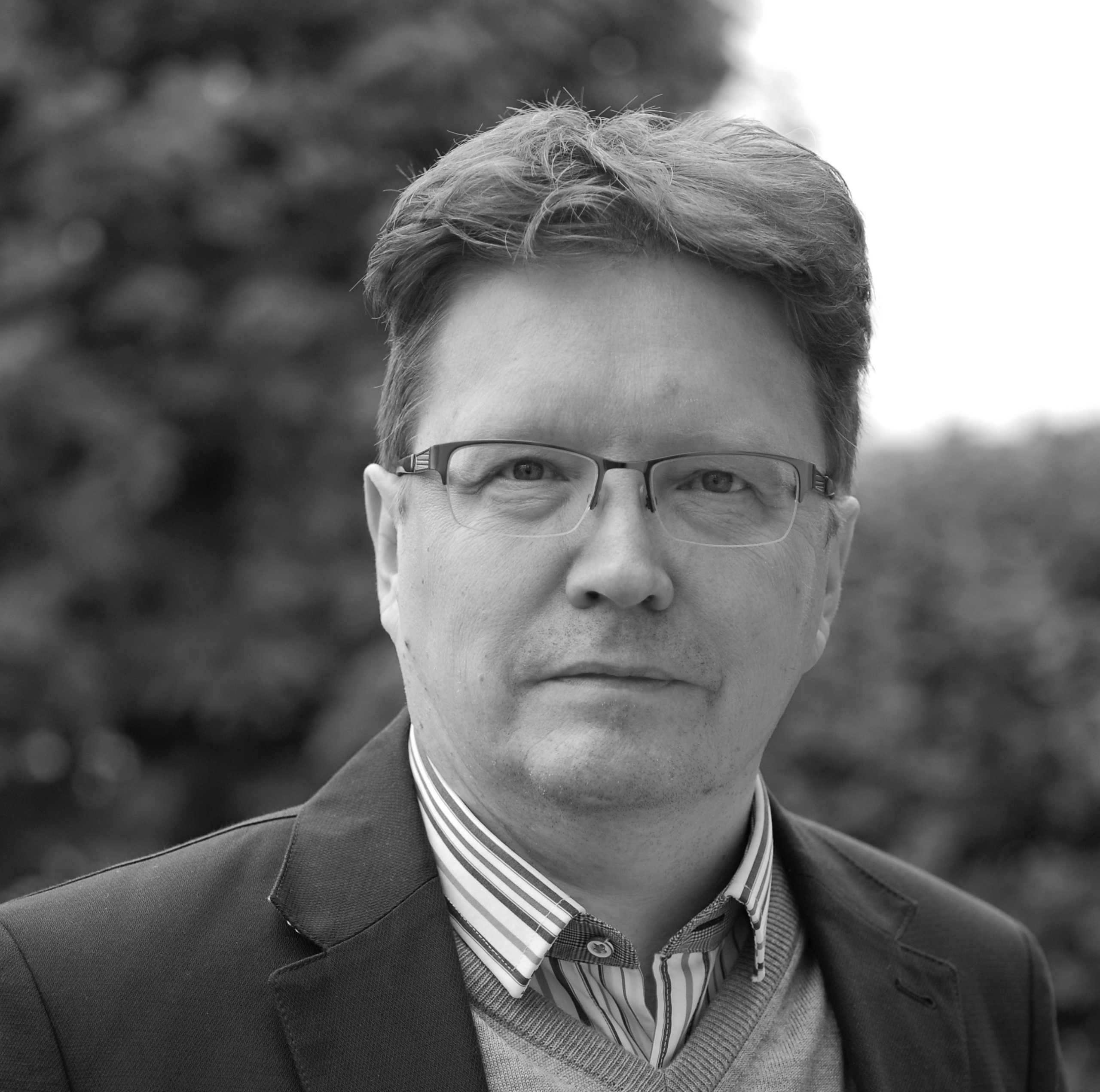 Mies, jolla on silmälasit, raitapaita, villapaita ja bleiseri, seisoo ulkona ja katsoo kameraan neutraalilla ilmeellä. Tausta on epätarkka, ja siinä näkyy puita tai pensaita. Kuva on mustavalkoinen.