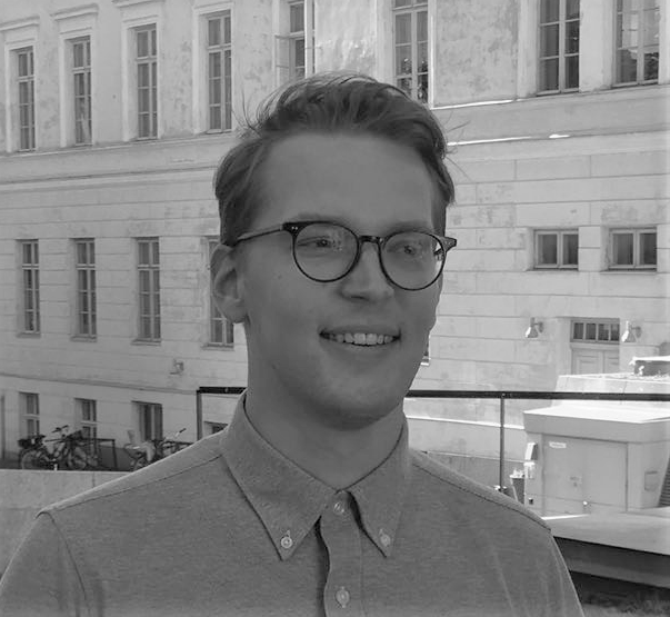 Nuori, silmälasipäinen ja lyhythiuksinen henkilö hymyilee seisoessaan ulkona suuren, vanhan rakennuksen edessä, jossa on paljon ikkunoita. Kuva on mustavalkoinen.