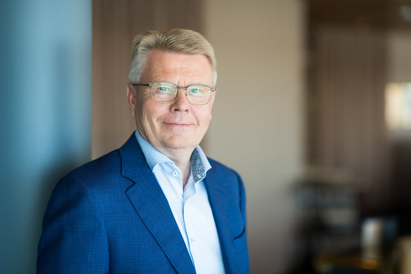Keski-ikäinen mies, jolla on lyhyet vaaleat hiukset ja silmälasit, sininen puvuntakki ja vaalea paita, seisoo sisätiloissa neutraalilla taustalla ja hymyilee hieman.