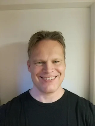 Lyhyet, vaaleanruskeat hiukset omaava mies hymyilee kameralle. Hänellä on yllään musta t-paita ja hän seisoo tavallisen, vaalean seinän edessä.