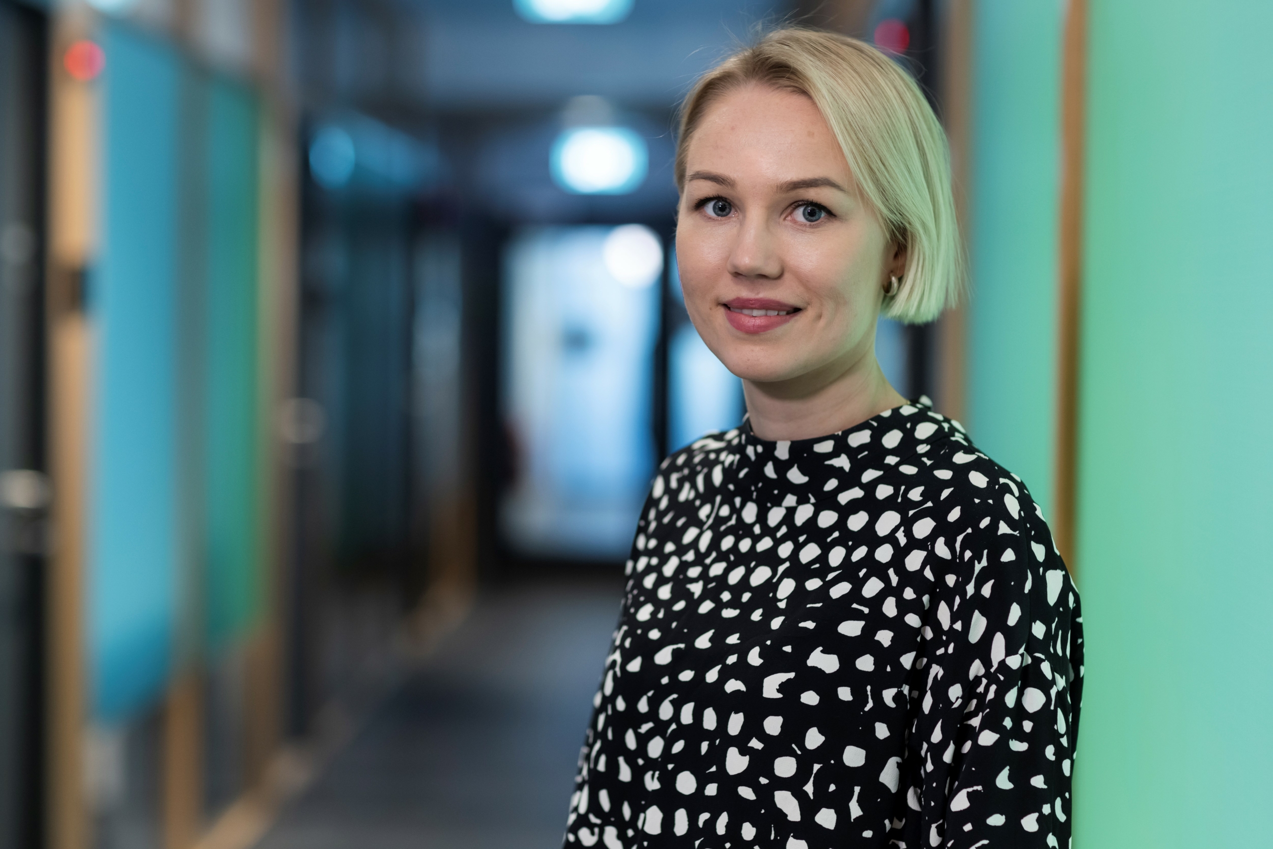 Lyhyet vaaleat hiukset omaava nainen, jolla on mustavalkoinen kuvioitu toppi, seisoo kirkkaasti valaistussa käytävässä, jonka seinät ovat tiilenpunaiset, ja hymyilee kameralle.