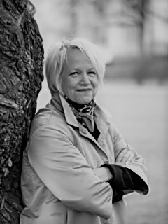 Lyhyet vaaleat hiukset omaava nainen seisoo ulkona puuta vasten nojaillen ja kädet ristissä. Hänellä on yllään vaalea takki ja huivi, ja hän hymyilee hieman. Tausta on sumea.